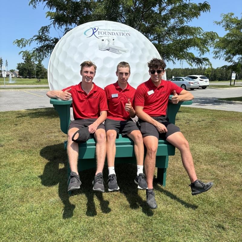 Three individuals in red shirts sitting on a large green bench with a visible logo in the background.