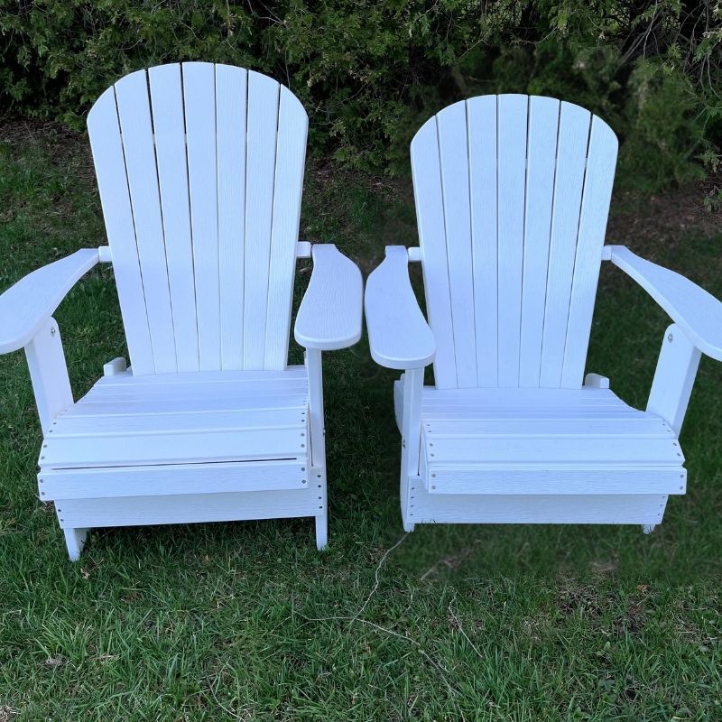 Two white Royal Upright Adirondack Chair, on a grassy area with greenery in the background.