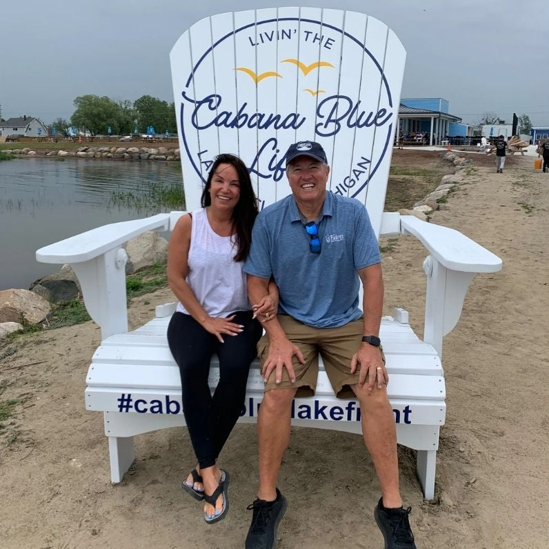 Two people sitting on Traditional Giant white chair with 'Living the Cabana Blue Life' branding by a lake.