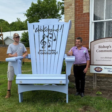 Two men standing next to Customized Giant blue chair with 'Music Builds Community!' text, in front of a brick building.