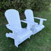 Two white Royal Upright Adirondack Chair, on grass with greenery in the background